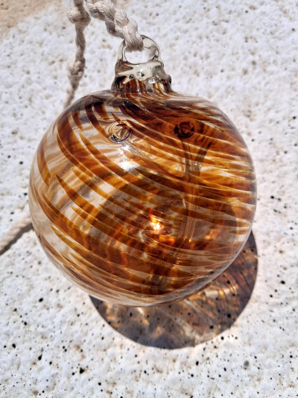 Amber swirl handblown glass witch ball with braided rope hanger resting on concrete surface with red brick wall background