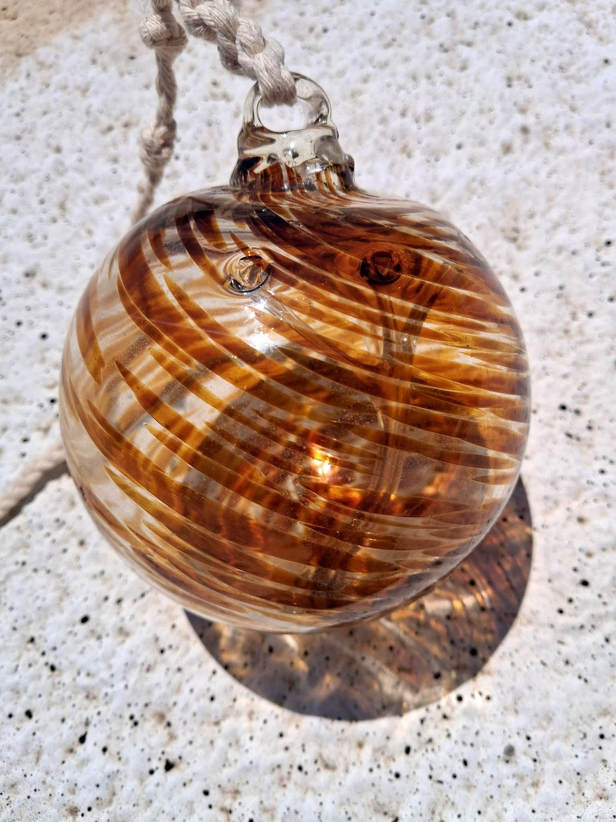 Amber swirl handblown glass witch ball with braided rope hanger resting on concrete surface with red brick wall background