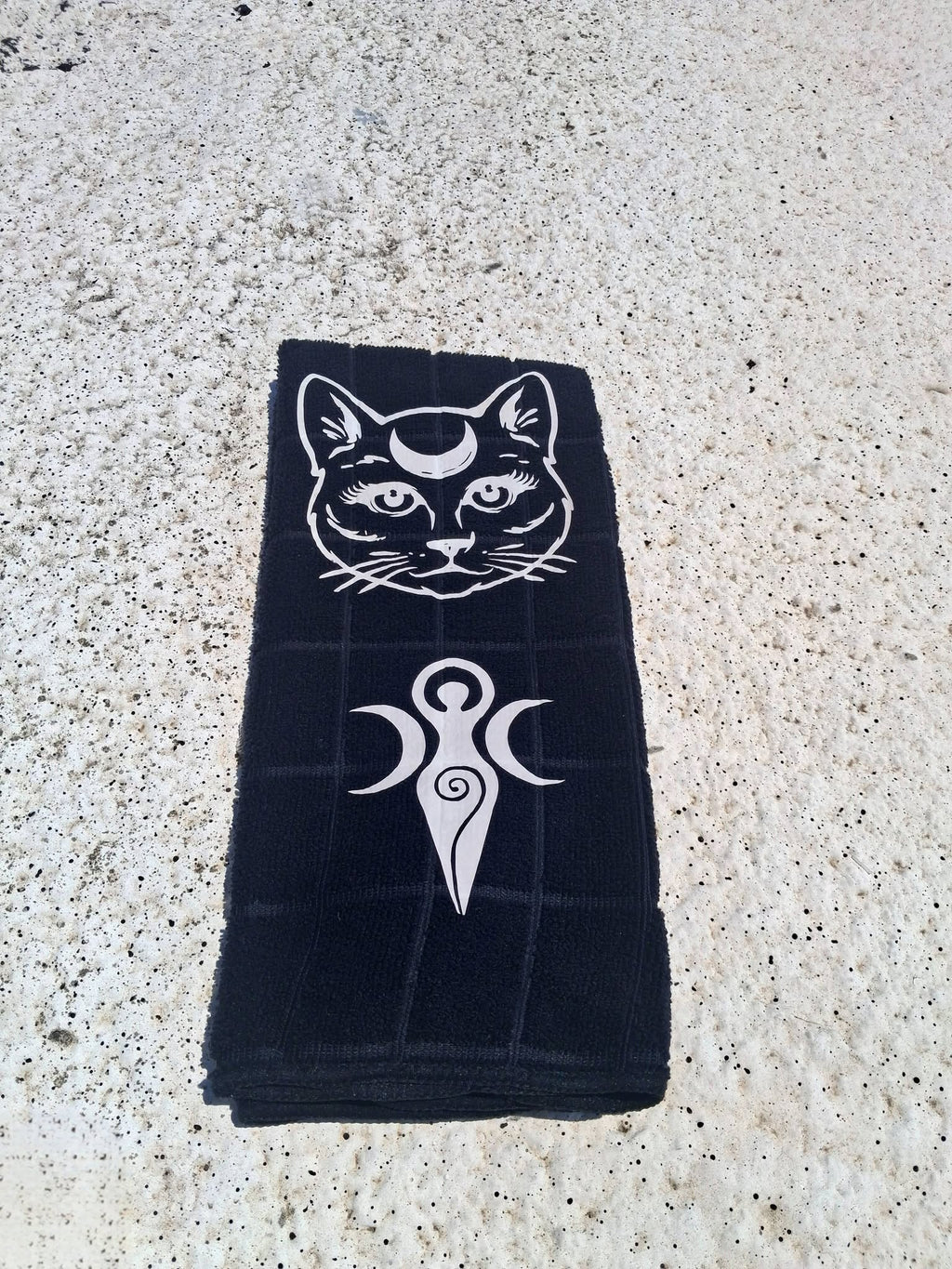 Black microfiber hand towel with white moon cat face and Triple Moon Goddess symbol handmade design folded on concrete